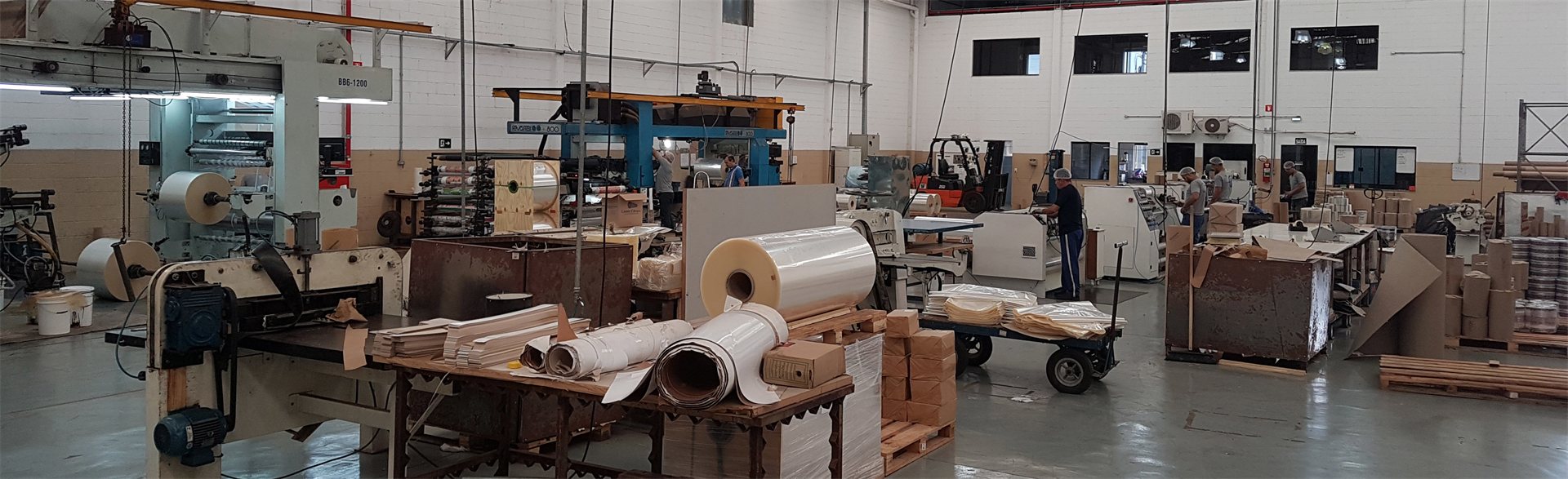 Vista panorâmica do chão de fábrica com impressora flexográfica, bobinas de filme e equipe em produção